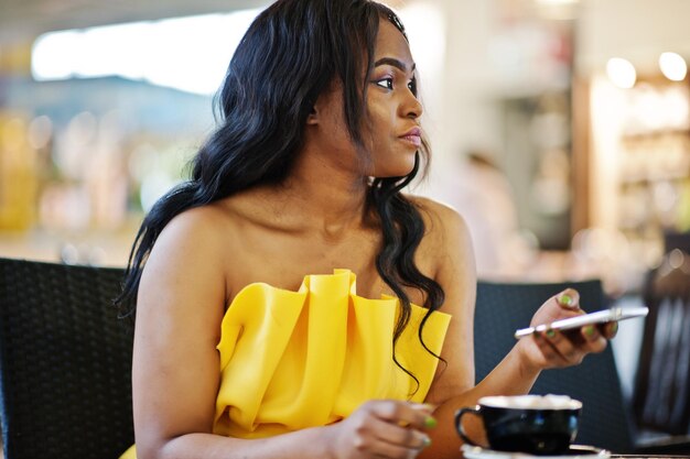 Mujer afroamericana con estilo en vestido amarillo posó en el café con una taza de capuchino y teléfono móvil en las manos