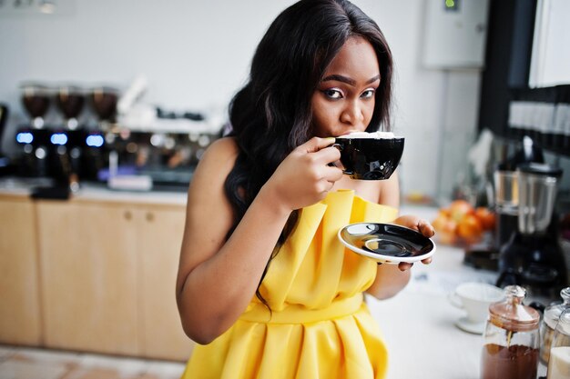 Mujer afroamericana con estilo en vestido amarillo posó en la cafetería con una taza de capuchino