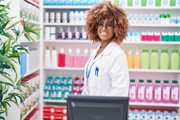 Mujer afroamericana farmacéutica sonriendo segura de pie en la farmacia