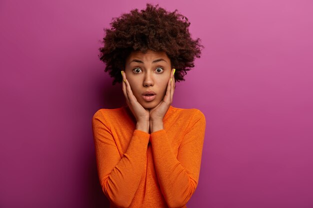 Mujer afroamericana fascinada agarra las mejillas, vestida con un jersey naranja, se da cuenta de que se olvidó de algo importante, recibe una gran multa, posa contra la pared violeta. Expresiones de rostro humano