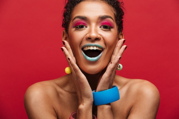 Mujer afroamericana feliz multicolor con maquillaje de moda gritando en la cámara tomados de la mano en la cara, sobre la pared roja