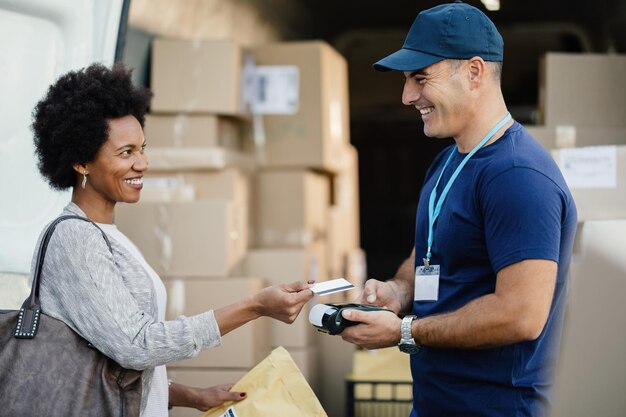 Mujer afroamericana feliz usando tarjeta de crédito mientras realiza un pago sin contacto a un mensajero para la entrega de paquetes