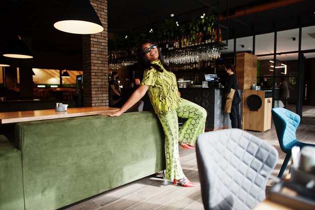 Mujer afroamericana glamorosa usa vestido de serpiente de moda y pose de gafas de sol en el restaurante