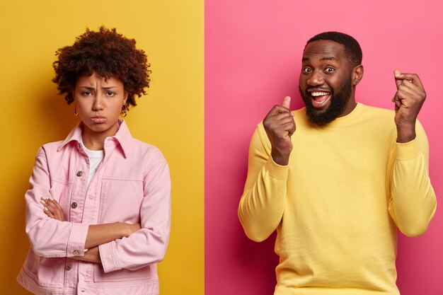 La mujer afroamericana insatisfecha se encuentra con los brazos cruzados, disgustada después de la pelea con el marido, el hombre de piel oscura triunfante levanta ambos brazos. Soporte de pareja étnica sobre pared de dos colores