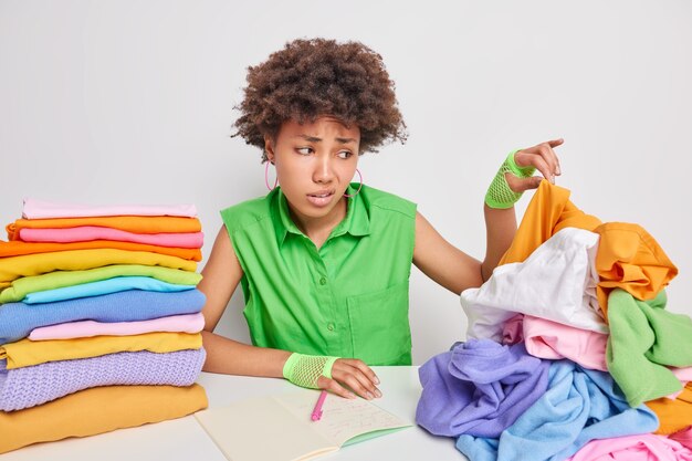 Mujer afroamericana insatisfecha recoge ropa sucia de trabajos de pila en la lavandería hace notas en poses de cuaderno contra la pared blanca siente aversión tiene cara de repugnancia. Gran concepto de blanqueo