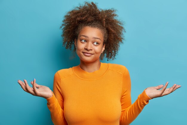 La mujer afroamericana joven desconcertada tiene el pelo rizado y tupido extiende las manos en la duda mira a un lado con la expresión de la cara inconsciente vestida con un jersey de manga larga naranja.