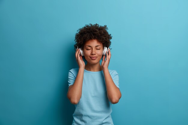 La mujer afroamericana joven rizada satisfecha tiene un estado de ánimo feliz y despreocupado, cierra los ojos y escucha música en auriculares, usa una camiseta azul informal, posa en el interior. Personas, ocio, concepto de entretenimiento.