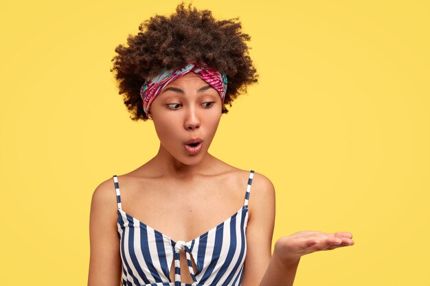 Mujer afroamericana joven sorprendida con cabello oscuro y rizado, vestida con ropa de verano a la moda, levanta la palma, finge sostener algo, mira con expresión de sorpresa