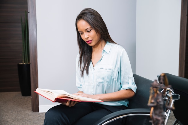 Mujer afroamericana con libro sentado en sillón en oficina