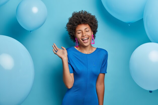 La mujer afroamericana llena de alegría cierra los ojos con alegría, usa un elegante vestido azul, modela sobre globos festivos, posa durante la celebración, tiene humor de fiesta La cumpleañera se mueve con ritmos de música