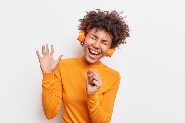 La mujer afroamericana llena de alegría mantiene la palma levantada tiene una expresión despreocupada, canta una canción, escucha música en auriculares, usa un jersey naranja casual aislado sobre una pared blanca. Entretenimiento divertido