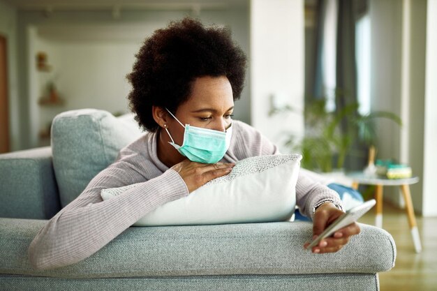 Mujer afroamericana con mascarilla usando teléfono móvil mientras descansa en el sofá de casa