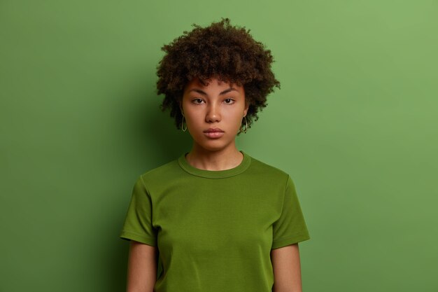 Una mujer afroamericana milenaria seria y decidida mira directamente, se ve confiada, usa una camiseta verde informal, posa en el interior, escucha información, va directo a los negocios