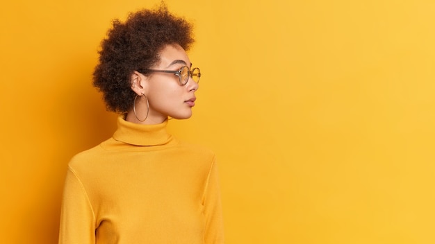 Mujer afroamericana mira a un lado con expresión pensativa gira la cabeza a un lado enfocada en la distancia, pensativamente, lleva gafas transparentes de cuello alto.