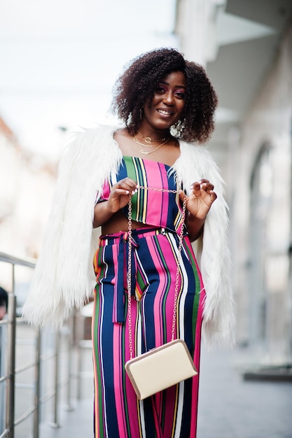 Mujer afroamericana de moda en mono rosa a rayas con abrigo de piel sintética esponjoso posado en la calle de megapolis