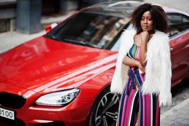 Mujer afroamericana de moda en mono rosa a rayas con abrigo de piel sintética esponjoso posado contra un coche de lujo rojo rico con teléfono móvil a mano