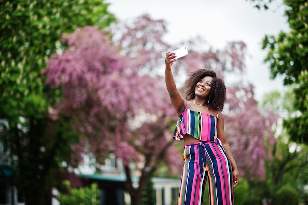 Mujer afroamericana de moda en traje de rayas rosas posado en la calle Spring Bloom y haciendo selfie por teléfono móvil