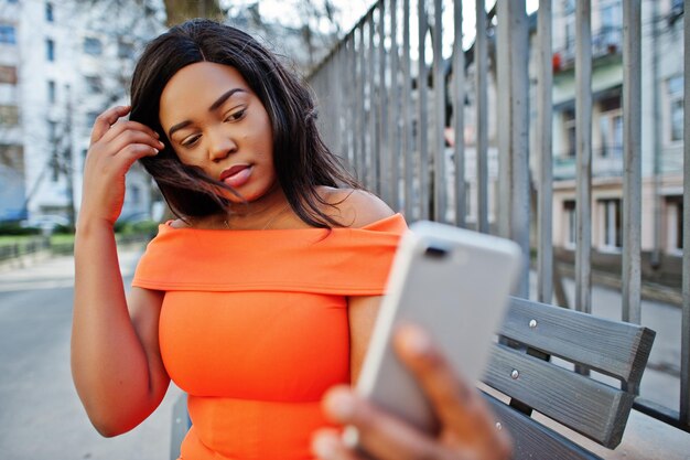 Mujer afroamericana modelo xxl en vestido naranja mirando en el teléfono móvil
