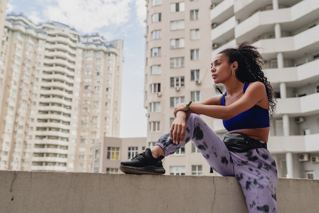 Foto gratuita mujer afroamericana negra en traje urbano de fitness deportivo en la azotea haciendo ejercicio escuchando música con auriculares