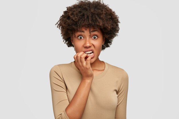 Mujer afroamericana nerviosa ansiosa muerde las uñas con expresión de desconcierto, tiene el pelo oscuro y rizado, vestida de manera informal, aislada sobre una pared blanca. ¡Dios mío, tengo miedo de esto! Concepto de emociones