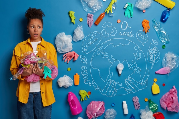 Foto gratuita mujer afroamericana nerviosa preocupada en camisa amarilla protege el medio ambiente de la basura, recoge basura, preocupada por la contaminación plástica, responsable de la limpieza del territorio.