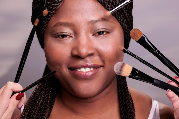 Mujer afroamericana obteniendo un retrato de maquillaje profesional. Las manos de los maquilladores aplican sombras de ojos y polvo en la cara del modelo, usando pinceles cosméticos decorativos de cerca
