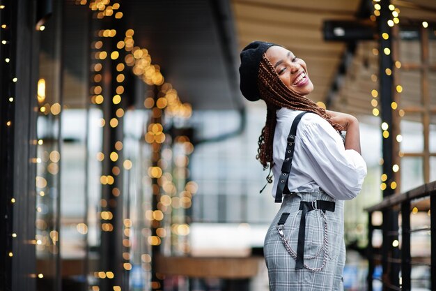 Mujer afroamericana en overol y boina posada en una terraza al aire libre con guirnaldas de adornos navideños