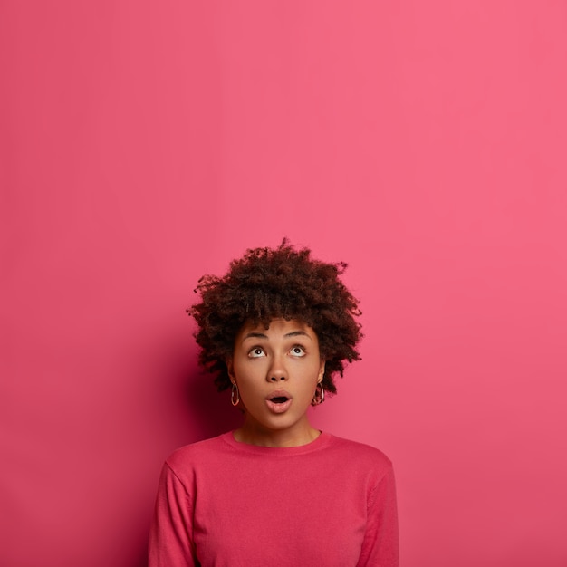 Foto gratuita mujer afroamericana sin palabras mantiene la boca ligeramente abierta, concentrada arriba, nota algo aterrador, vestida con ropa casual, posa sobre una pared rosa, espacio libre para su anuncio