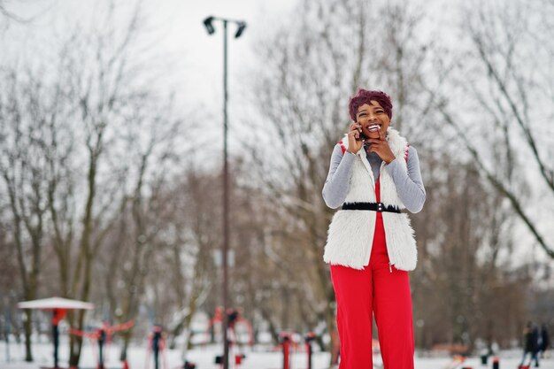 Mujer afroamericana con pantalones rojos y chaqueta de abrigo de piel blanca posada en el día de invierno contra un fondo nevado hablando por teléfono