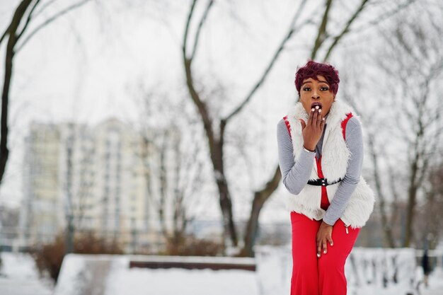 Mujer afroamericana con pantalones rojos y chaqueta de abrigo de piel blanca posada en el día de invierno contra un fondo nevado muestra una emoción sorprendida