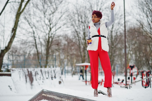 Mujer afroamericana con pantalones rojos y chaqueta de abrigo de piel blanca posada en el día de invierno contra un fondo nevado y sosteniendo una taza de café