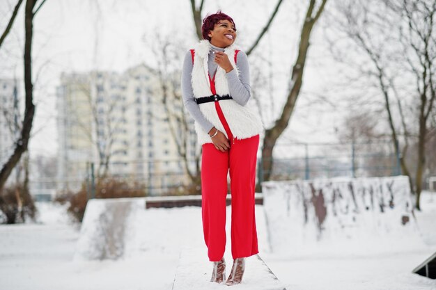 Mujer afroamericana con pantalones rojos y chaqueta de abrigo de piel blanca posada en el día de invierno contra un fondo nevado