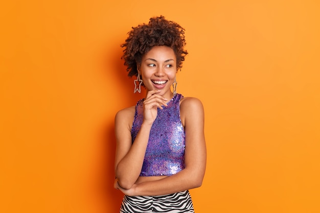 Foto gratuita la mujer afroamericana de pelo rizado de moda atractiva en ropa elegante sostiene la barbilla mira hacia otro lado con alegría tiene poses de humor positivo contra vestidos de fondo naranja para fiesta o cita. concepto de estilo