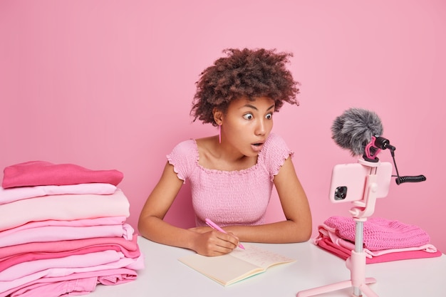 Mujer afroamericana de pelo rizado sorprendida toma notas en un cuaderno enfocado en la cámara del teléfono inteligente se sienta en la mesa rodeada de pilas de ropa cuidadosamente doblada hace video blog sobre la rutina doméstica