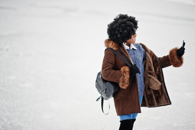 Mujer afroamericana de pelo rizado vestida con abrigo de piel de oveja y guantes con mochila posada en el día de invierno