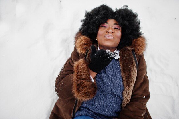 Mujer afroamericana de pelo rizado vestida con abrigo de piel de oveja y guantes posados en el día de invierno yace en la nieve