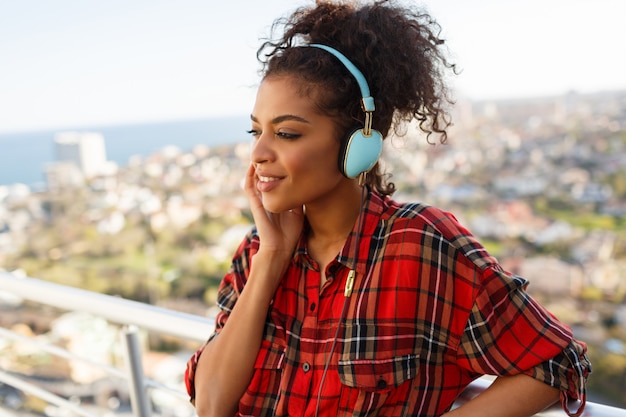Mujer afroamericana con piel oscura posando con auriculares en la azotea. Fondo de paisaje urbano