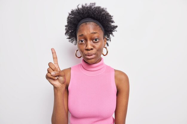 Mujer afroamericana de piel oscura sorprendida con cabello rizado natural apunta el dedo índice hacia arriba muestra algo