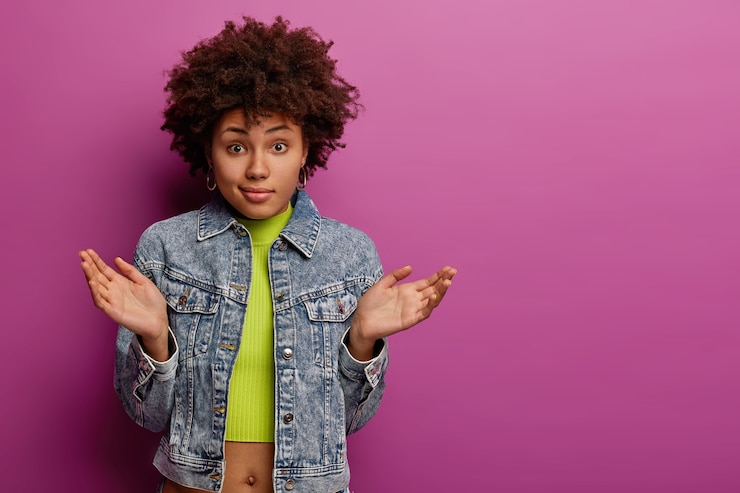 Foto gratuito una mujer afroamericana sin pistas y dudosa extiende las palmas de las manos con vacilación, no puede tomar una decisión, usa una chaqueta vaquera y un top, tiene una mirada desconcertada, posa sobre una pared violeta vibrante, copia el área del espacio a un lado