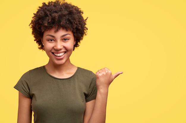 La mujer afroamericana positiva de aspecto agradable y divertida señala a la derecha, vestida con una camiseta informal, muestra el espacio libre contra la pared amarilla, sugiere ir allí, tiene amigos