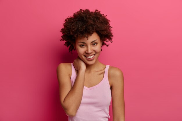 La mujer afroamericana positiva complacida mira directamente con una sonrisa encantadora, toca el cuello suavemente, se siente satisfecha, tiene el pelo rizado y tupido, vestida con ropa casual, aislada en la pared rosa