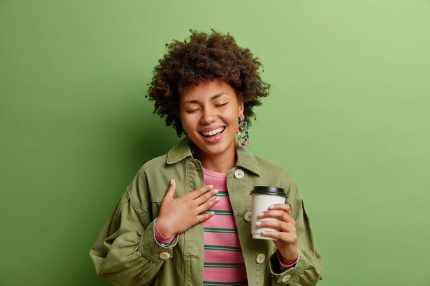 Mujer afroamericana positiva eith cabello rizado cierra los ojos de la felicidad mantiene la mano cerca del corazón bebe café para llevar disfruta del tiempo libre vestida con ropa de moda aislada en la pared verde