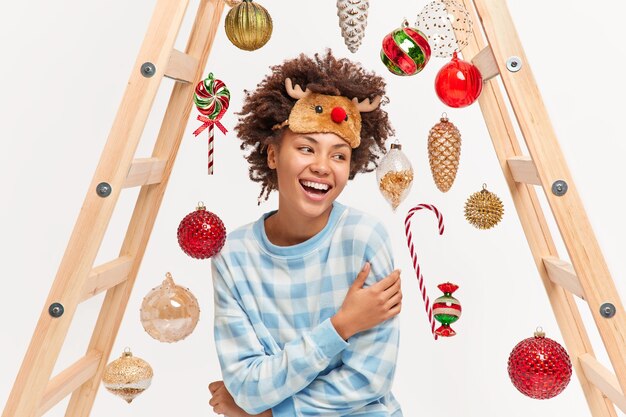 Una mujer afroamericana positiva está llena de sonrisas de felicidad y muestra unos dientes perfectos blancos, usa ropa de dormir cómoda, mira poses a un lado