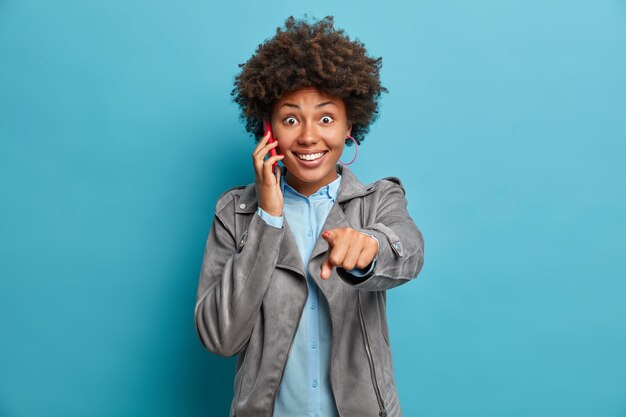 Una mujer afroamericana positiva indica directamente a la cámara que tiene una conversación telefónica y sonríe ampliamente