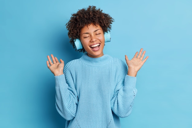 La mujer afroamericana positiva con el pelo rizado levanta las palmas de las manos se divierte mientras escucha la pista de audio lleva auriculares inalámbricos vestidos con un jersey informal