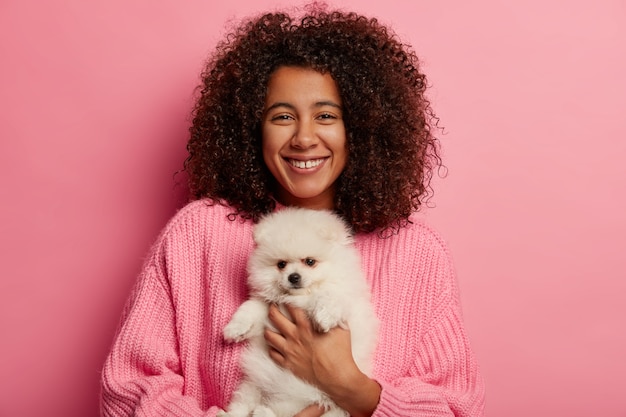 La mujer afroamericana positiva posa con spitz esponjoso en las manos, acaricia a un perro, tiene una expresión alegre para adoptar un animal doméstico aislado sobre fondo rosa.