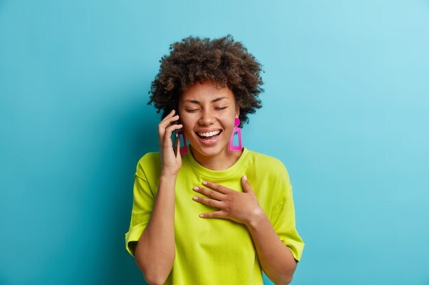 Mujer afroamericana positiva relajada despreocupada se ríe mientras habla a través del teléfono inteligente mantiene la mano en el pecho cierra los ojos expresa emociones positivas oye una broma divertida vestida informalmente se encuentra en el interior