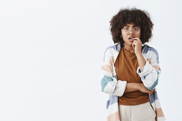 Mujer afroamericana preocupada, pensativa e inteligente curiosa con peinado afro en vasos transparentes mordiéndose la uña y frunciendo el ceño mientras piensa cómo tomar una decisión difícil