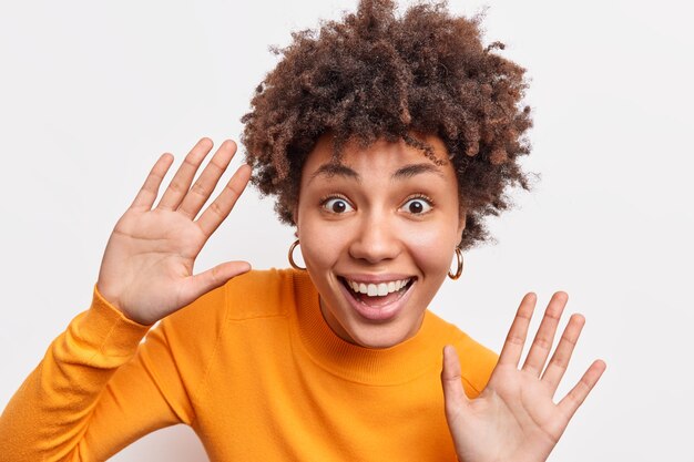 La mujer afroamericana rizada juguetona expresiva sonríe ampliamente levanta las palmas de las manos tontas alrededor tiene buen humor usa un jersey naranja aislado sobre una pared blanca se siente emocionado expresa alegría