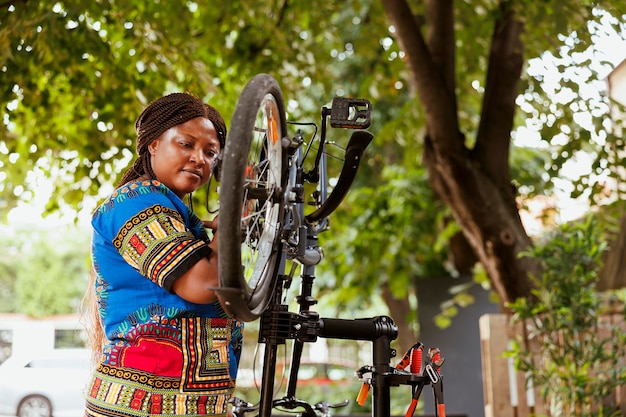 Mujer afroamericana sana enfocada reparando su propia bicicleta en el patio y realizando mantenimiento anual al aire libre usando herramientas expertas. La ciclista femenina inspecciona y repara los pedales de las bicicletas.